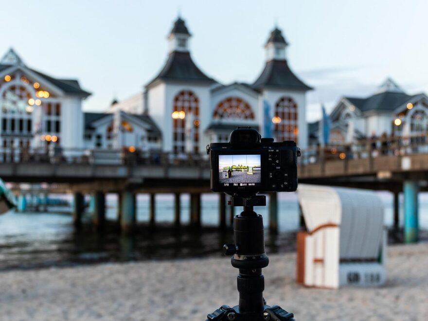 Reisestativ mit Kamera am Strand, im Hintergrund eine Seebrücke.