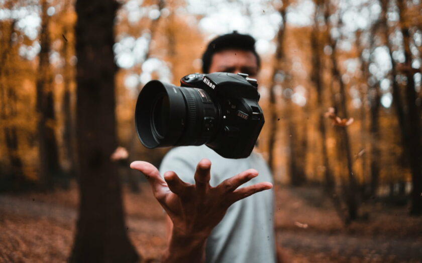Person wirft eine Kamera in einem herbstlichen Wald in die Luft, symbolisiert kreative Fotografie.