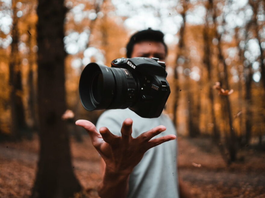 Person wirft eine Kamera in einem herbstlichen Wald in die Luft, symbolisiert kreative Fotografie.
