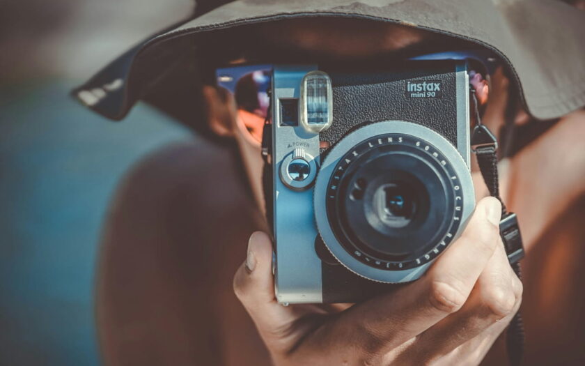 Person mit braunem Fischerhut hält eine schwarze und graue Fujifilm Instax Sofortbildkamera, bereit, ein Foto aufzunehmen.