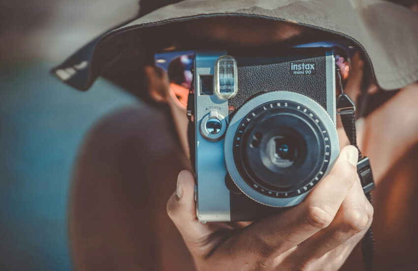Person mit braunem Fischerhut hält eine schwarze und graue Fujifilm Instax Sofortbildkamera, bereit, ein Foto aufzunehmen.