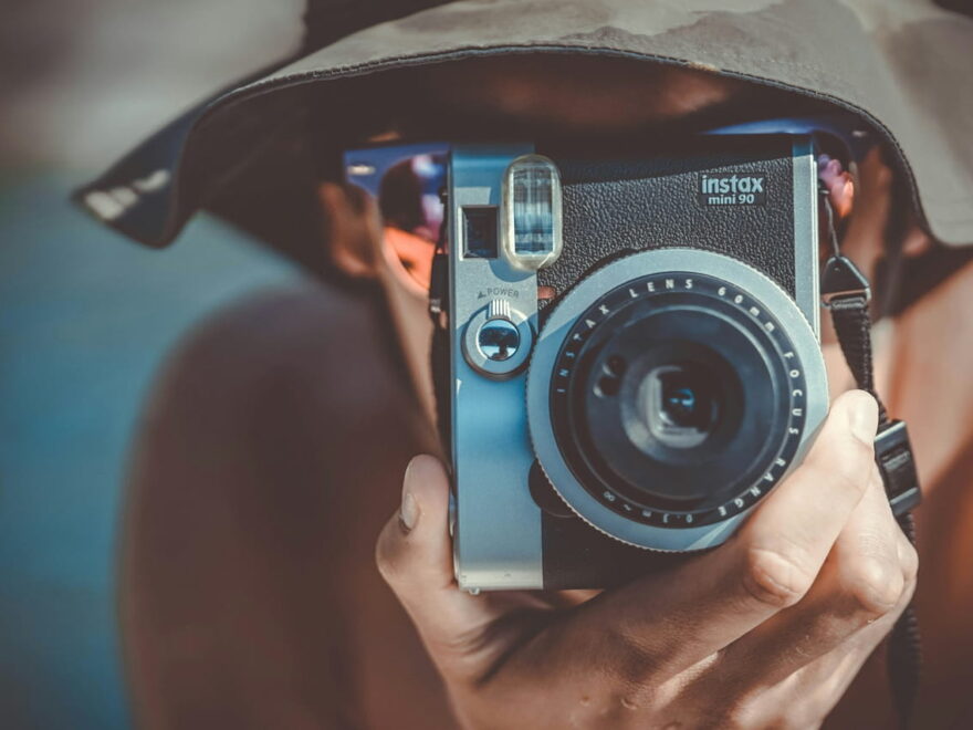 Person mit braunem Fischerhut hält eine schwarze und graue Fujifilm Instax Sofortbildkamera, bereit, ein Foto aufzunehmen.