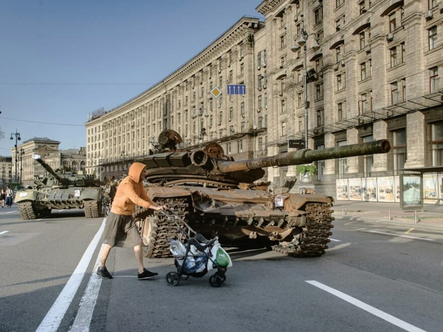 Person schiebt Kinderwagen vorbei an zerstörten Panzern auf einer städtischen Straße, Symbol für Reportagefotografie in Kriegs- und Krisengebieten.